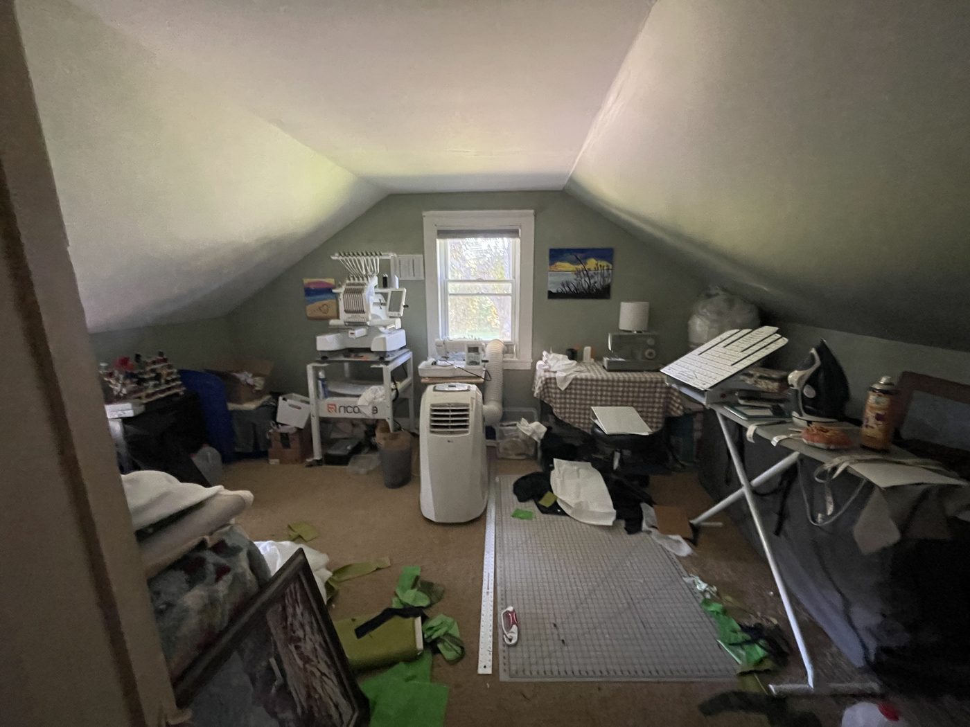 The guest and craft room before the work started: a sloped-ceiling room with sage green walls, a multi-needle embroidery machine on a rolling stand beside the window, a sewing machine, an ironing board, a cutting mat on the carpet, and fabric offcuts, boxes and half-finished projects across the floor.