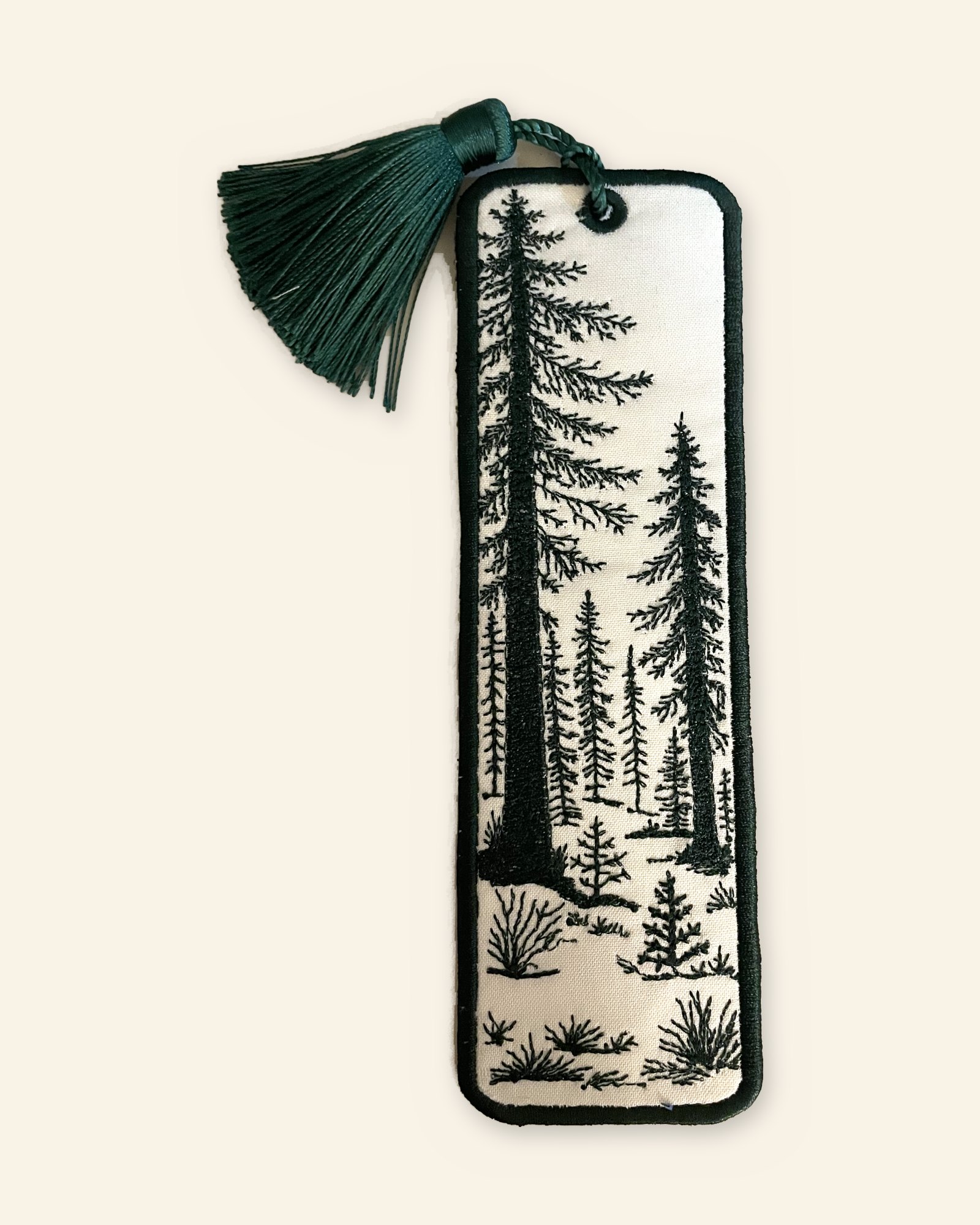 The In the Pines bookmark: tall pine trees and brush embroidered in dark green on a cream ground, with a green tassel at the top.