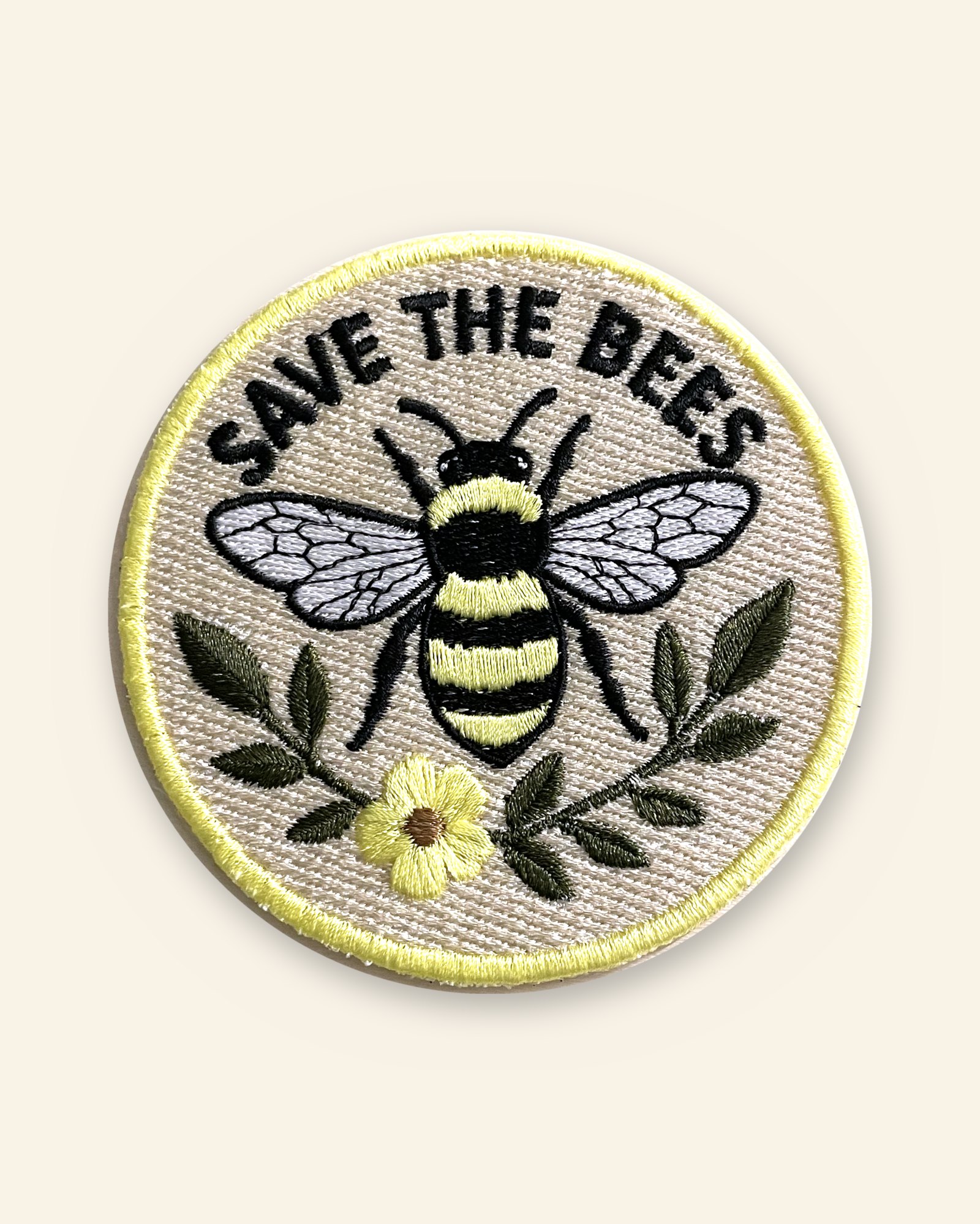 The Save the Bees patch: a round embroidered patch with a black and yellow bumblebee, grey veined wings, green leaves and a yellow flower on a cream twill ground, the words Save the Bees arched over the top, and a yellow border.